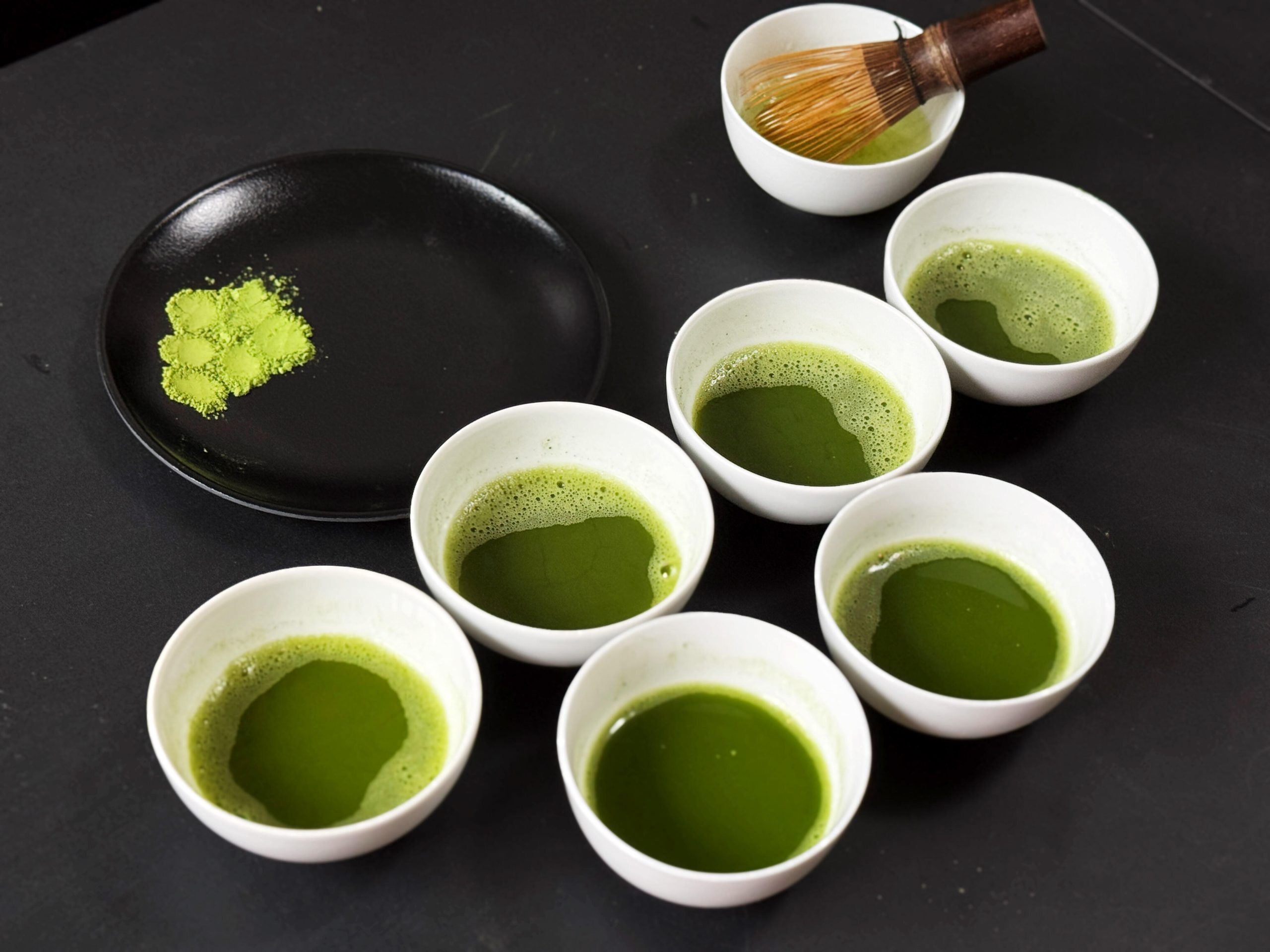 Whisked matcha review samples in white bowls beside a powder plate and bamboo whisk