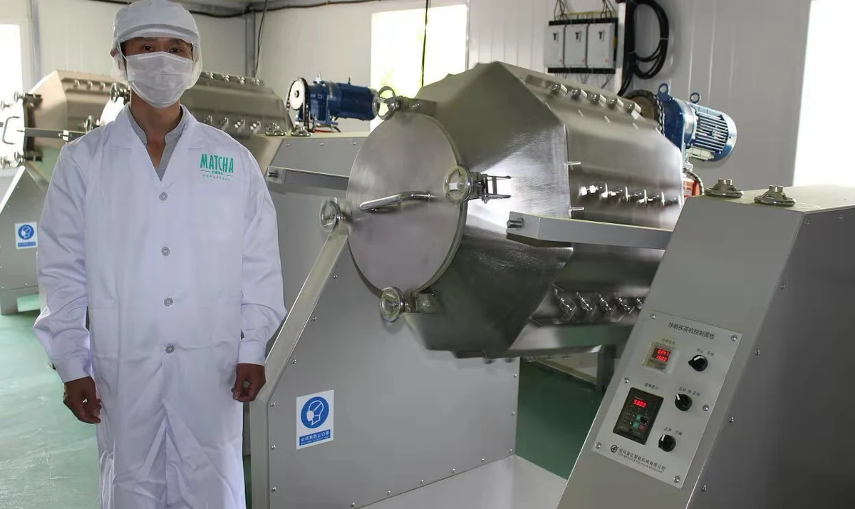 Production team member wearing a cap and mask beside enclosed processing equipment
